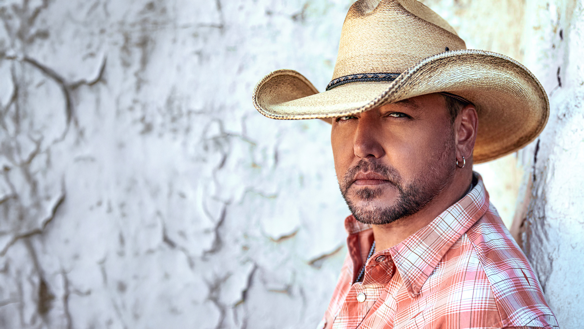 Country artist Jason Aldean dressed in a collared shirt, posing for the camera against a wall.