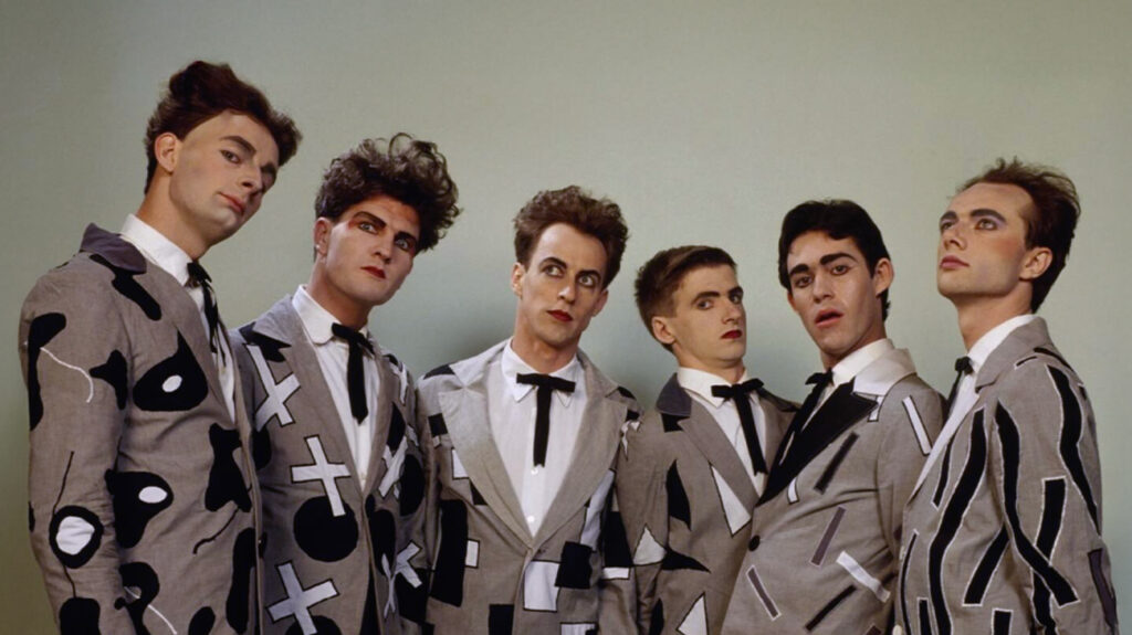 Members of the band Split Enz, dressed in suits, standing together and smiling for a group photo.