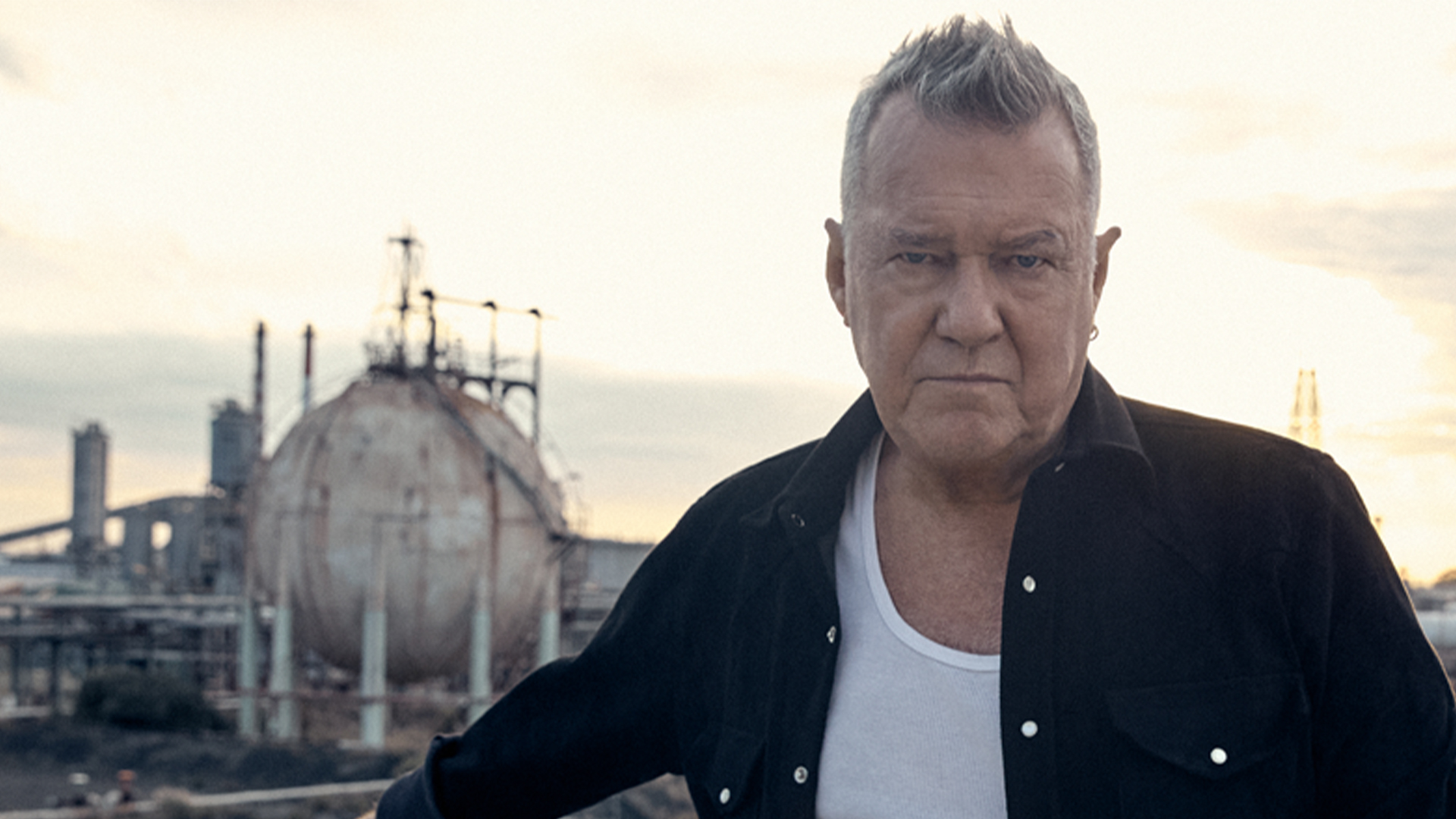 Singer Jimmy Barnes dressed in casual clothing posing against an industrial background.