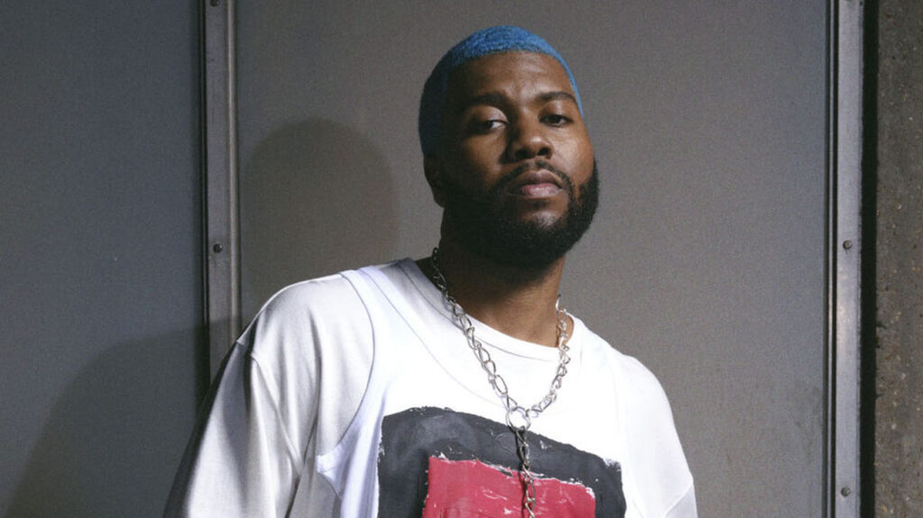 Khalid with a blue beard wearing a white shirt, smiling and looking directly at the camera.