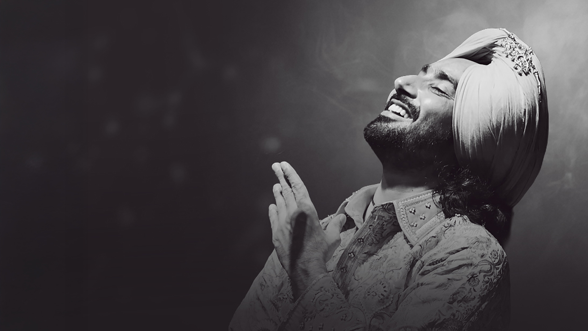 Satinder Sartaaj with hands-clapped and a smiling pose against a grey background.
