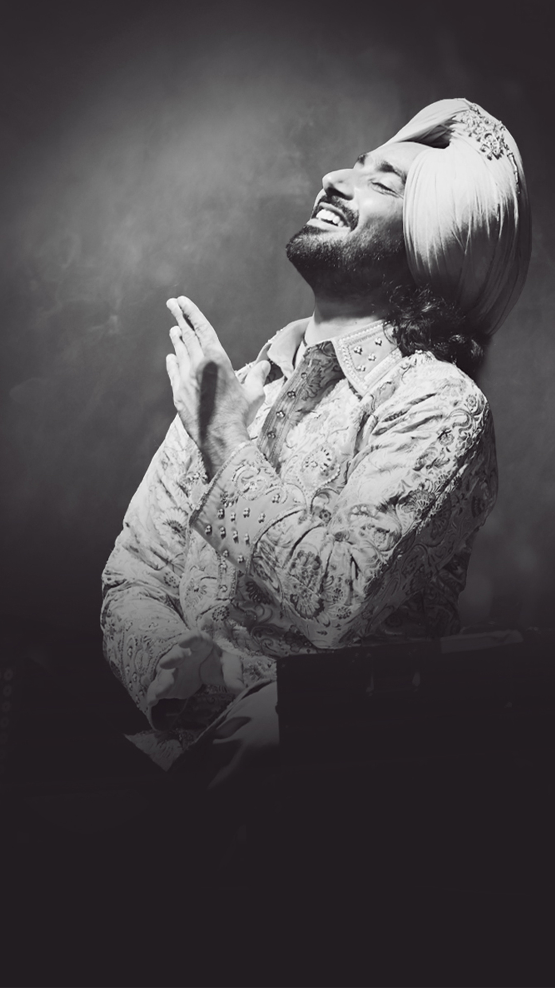 Satinder Sartaaj with hands-clapped and a smiling pose against a grey background.
