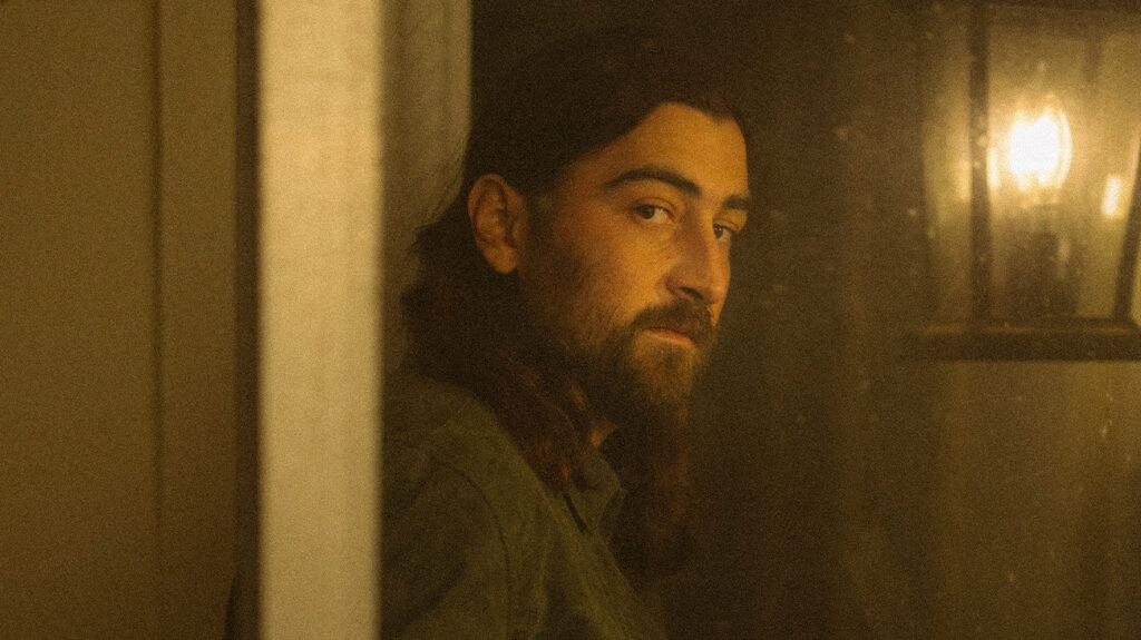 Noah Kahan, with long hair and a beard, looks pensively out the window.