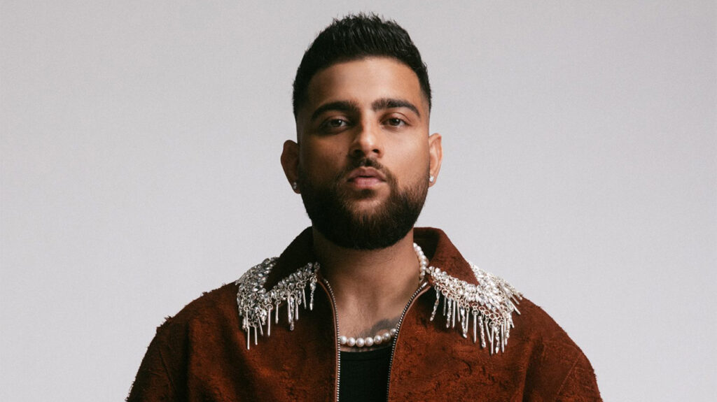 Karan Aujla wearing a decorative necklace and jacket posing against a neutral background.