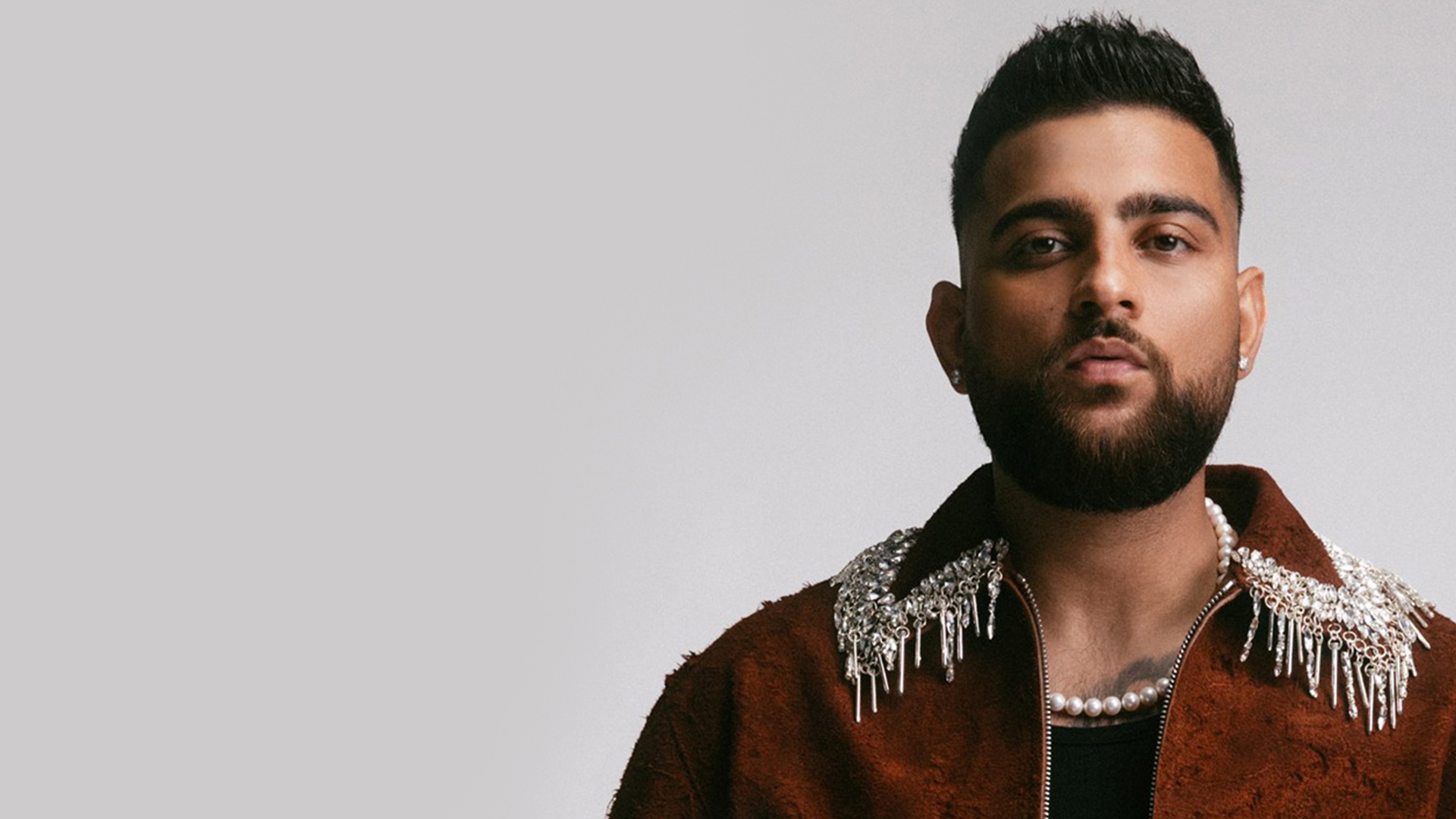 Karan Aujla wearing a decorative necklace and jacket posing against a neutral background.