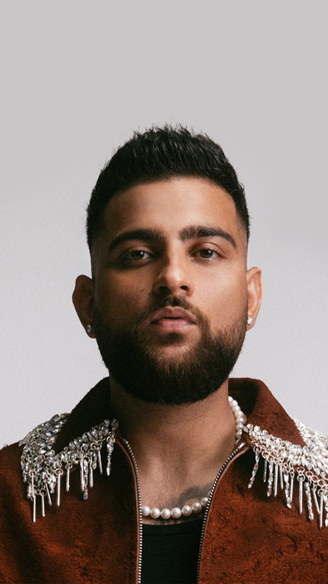 Karan Aujla wearing a decorative necklace and jacket posing against a neutral background.